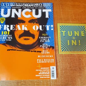 May include: A magazine cover with the title "UNCUT" and the words "Freak Out!" in large, bold letters. The cover features a photo of a man with a mustache and beard. The magazine is yellow and orange with black text. The magazine is next to a smaller yellow and blue card with the words "Tune In!" in large, bold letters.