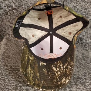 Peut inclure: Une casquette de baseball camouflage avec une visi&egrave;re noire et un dos en maille blanche. L'int&eacute;rieur de la casquette est blanc avec des coutures noires et un petit logo orange. La casquette a une fermeture velcro.