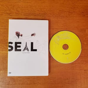 May include: A DVD case and a CD are on a wooden surface. The DVD case is white with the word "SEAL" in large black letters, a drawing of a person, and the Eiffel Tower. The CD is yellow with scribbled lines.