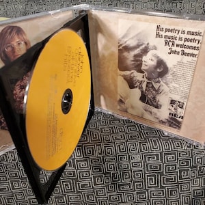 May include: A John Denver Greatest Hits CD in a clear plastic jewel case. The disc is yellow with black text. The insert features a black and white photograph of John Denver playing guitar with the text His poetry is music. His music is poetry. RCA welcomes John Denver.