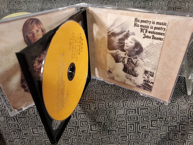 May include: A John Denver Greatest Hits CD in a clear plastic jewel case. The disc is yellow with black text. The insert features a black and white photograph of John Denver playing guitar with the text His poetry is music. His music is poetry. RCA welcomes John Denver.
