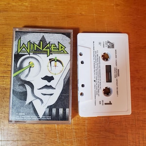 May include: A Winger cassette tape with a black, white, and green album cover featuring a stylized face with crosshairs. The white cassette tape has the track list and "1" printed on it.