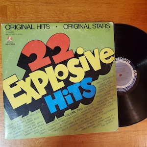 May include: A vintage vinyl record album titled "22 Explosive Hits" with a colorful cover featuring bold text and a list of song titles. The record is black and sits next to the album cover on a wooden surface.
