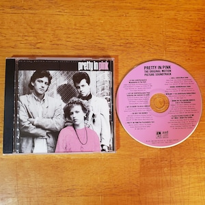 May include: A Pretty in Pink original motion picture soundtrack CD in a plastic case with a black spine. The cover features a black and white photo of three people. The disc is pink with black text listing track titles and production credits.