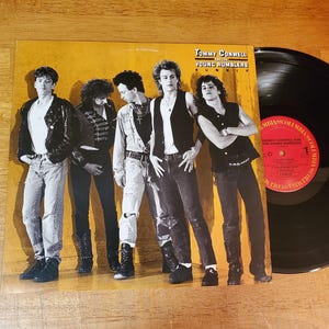 May include: Album cover for "Tommy Conwell and the Young Rumblers" featuring a band of five men in various outfits. The album is on a wooden surface with a vinyl record visible.