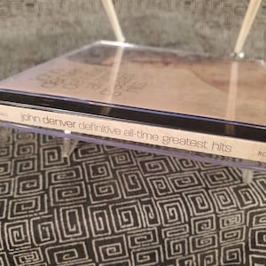 May include: A clear plastic CD jewel case containing the John Denver Definitive All-Time Greatest Hits album. The spine features the text John Denver Definitive All-Time Greatest Hits and the RCA/BMG logo. The cover art is visible through the case.