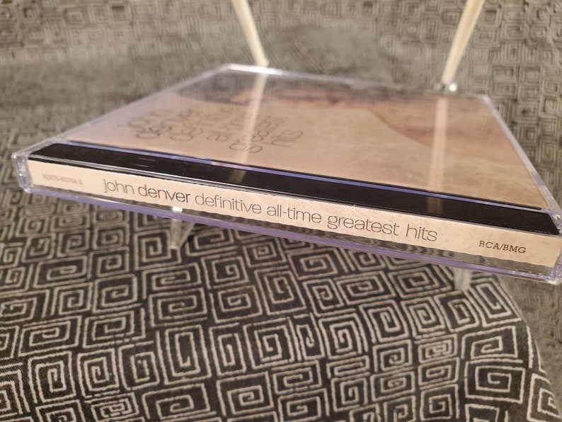 May include: A clear plastic CD jewel case containing the John Denver Definitive All-Time Greatest Hits album. The spine features the text John Denver Definitive All-Time Greatest Hits and the RCA/BMG logo. The cover art is visible through the case.
