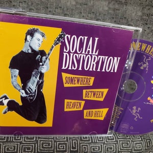 May include: A Social Distortion CD album titled "Somewhere Between Heaven and Hell." The album cover features a man playing a guitar against a yellow background, with the band's name and album title in bold text. The CD is visible.