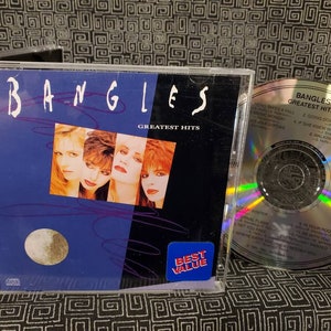 May include: A CD case with a blue background featuring a photo of four women. The title "BANGLES GREATEST HITS" is printed on the front of the case. The CD is visible in the background with a rainbow holographic effect.