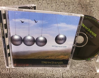 May include: A CD case with a black and white image of a Newton's cradle with three silver balls. The background is a blue sky with two birds flying. The text "Dream Theater" and "Octavarium" are printed on the case.