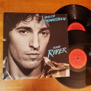 May include: Bruce Springsteen's album "The River" on vinyl. The album cover features a black and white portrait of Springsteen with the album title in blue. Two black vinyl records are visible.