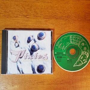 May include: A compact disc and its case are displayed on a wooden surface. The CD case features the band name "Pixies" in a stylized font, with the album title "Trompe Le Monde." The CD itself is green with white graphics.