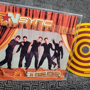 May include: A CD case featuring the band *NSYNC. The album cover shows the band members as puppets with the text "N SYNC" and "NO STRINGS ATTACHED". A second CD is visible with a yellow and brown circular design.