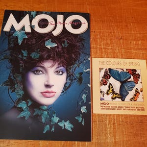 May include: A magazine cover featuring a woman with dark hair and blue eyes, surrounded by green leaves. The magazine is titled "MOJO The Music Magazine". A smaller card is next to the magazine, advertising a compilation album titled "The Colours of Spring" with a list of artists including The Weather Station, Sonnie Prince, Billy Bragg, Django Reinhardt, Mighty  Sake, Real Estate and more.
