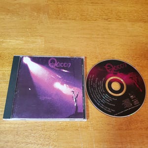 May include: A Queen CD album with the band's logo and album art. The CD case is black, and the disc is visible next to it. The album art features a silhouette of a person with a spotlight and the word "Queen".