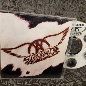 May include: Aerosmith's "Get a Grip" CD in a clear jewel case. The album cover features the band's logo with wings over a cowhide pattern. The CD is visible, displaying the band's logo and track details. The spine reads "Aerosmith Get a Grip".