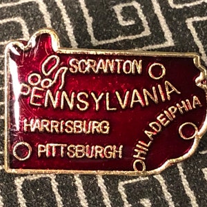 May include: A red enamel pin shaped like the state of Pennsylvania with the words "Pennsylvania", "Scranton", "Harrisburg", "Pittsburgh", and "Philadelphia" written in gold.