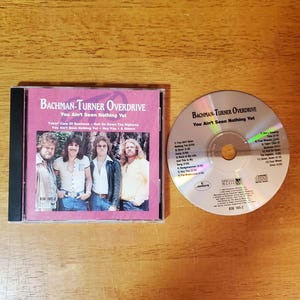 May include: A CD and its case featuring the album "You Ain't Seen Nothing Yet" by Bachman-Turner Overdrive. The CD case is pink with a photo of the band. The CD is silver with black text.