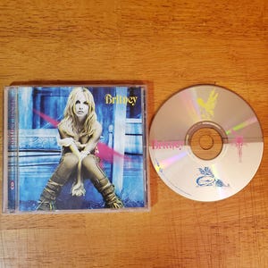 May include: A CD case and disc featuring Britney Spears. The CD case has a blue and white background with a photo of Britney Spears. The CD has a silver surface with a pink and yellow design.