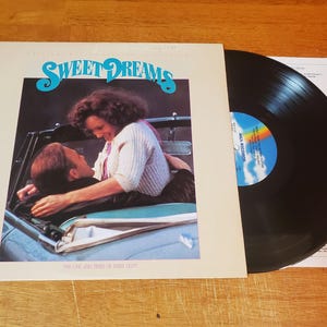 May include: An album cover for "Sweet Dreams" featuring a couple in a car, with the album title in turquoise script. A black vinyl record is next to the cover, with a rainbow label. The album is "The Life and Times of Buddy Holly."