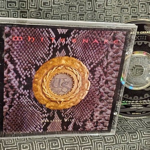 May include: A CD case with a snake skin pattern and a gold coin with the letters "US" on it. The title "Whitesnake's Greatest Hits" is printed on the side of the case. The CD is titled "Greatest Hits" and features a snake design.