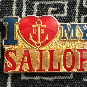 May include: A gold-colored pin with the words "I Love My Sailor" in red and blue letters. A red heart with a gold anchor and the letters "US" is in the center of the pin.