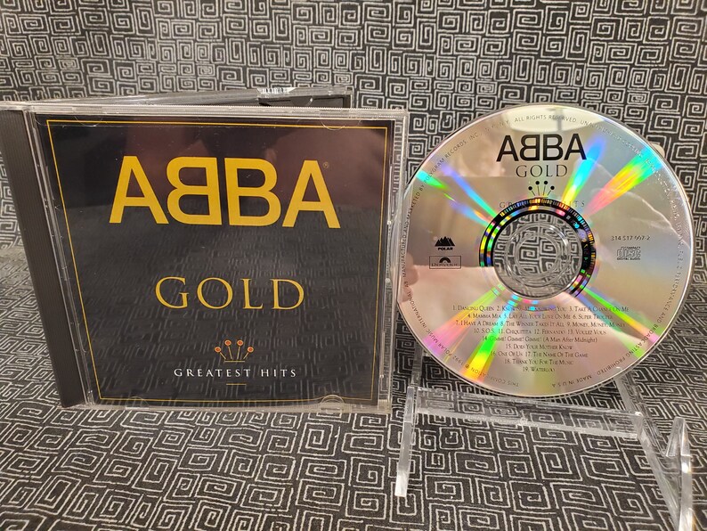 May include: ABBA Gold CD, a greatest hits compilation. The CD case is black with gold lettering and a gold crown design. The CD itself is silver with the ABBA logo and song titles visible. The CD is displayed on a clear plastic stand.