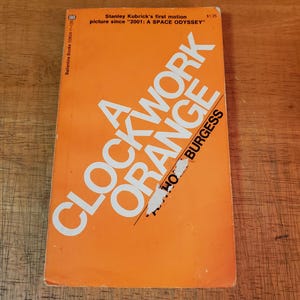 May include: A vintage paperback book with an orange cover. The title "A Clockwork Orange" is displayed in large white letters. The author's name, Burgess, is also visible. The cover includes text about Stanley Kubrick's film.