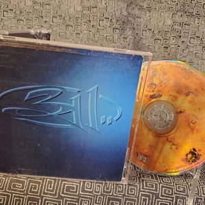 May include: A CD case with the band name "311" in a stylized font. The case has a blue gradient background. The CD is visible and has a gold and brown design.