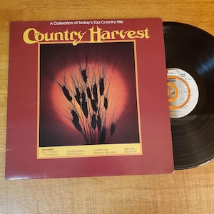 May include: A vinyl record album titled Country Harvest, featuring a maroon cover with a central image of wheat stalks against a red sun. The text includes A Collection of Today's Top Country Hits and lists artists like Billy Crash Craddock.