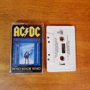 May include: A vintage AC/DC "Who Made Who" cassette tape. The cassette case features the band's logo and album art with a figure playing a guitar. The white cassette tape is next to the case. The album title and other text are visible.