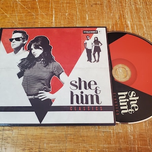 May include: A red, white, and black CD case with a red and black CD. The case features a photo of a man and woman, and the text "She & Him Classics".