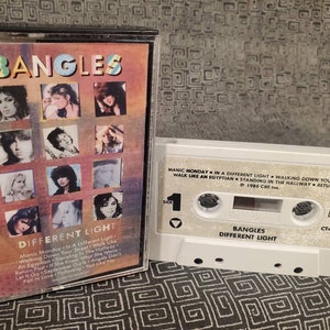 May include: A vintage cassette tape of The Bangles' album "Different Light." The album cover features a collage of images of the band members. The cassette is white with song titles and album information printed on it. The album was released in 1986.