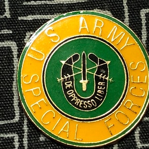May include: A gold and green pin with the words "US Army Special Forces" around the edge. The center of the pin features a green circle with a black horseshoe, crossed arrows, and a sword. The text "De Opresso Liber" is written inside the horseshoe.