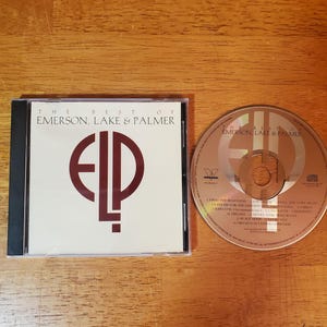 May include: A CD case and disc featuring "The Best of Emerson, Lake & Palmer." The CD case is white with a black spine and the letters "ELP" in a maroon color. The CD is brown with the band's name and song titles.