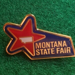 May include: A gold-colored pin with a red star and a blue background. The star has a white outline and a white silhouette of the state of Montana inside. The text "MONTANA STATE FAIR" is printed in gold letters on the blue background.