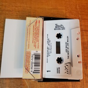 K&ouml;nnte beinhalten: Eine wei&szlig;e Kassette mit dem Text "JOE WALSH THE BEST OF JOE WALSH" und einem schwarzen Kassettenetui. Auf der Kassette steht "ROCKY MOUNTAIN WAY" und "TURN TO STONE". Die Kassette liegt auf einer Holzoberfl&auml;che.