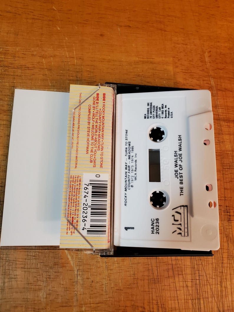 K&ouml;nnte beinhalten: Eine wei&szlig;e Kassette mit dem Text "JOE WALSH THE BEST OF JOE WALSH" und einem schwarzen Kassettenetui. Auf der Kassette steht "ROCKY MOUNTAIN WAY" und "TURN TO STONE". Die Kassette liegt auf einer Holzoberfl&auml;che.