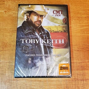 May include: DVD case featuring Toby Keith with the text "Toby Keith" in white capital letters. The DVD case has the CMT Pick logo and states "Features Programming From CMT" and "Over 2 Hours of DVD Content". The image is on a wooden surface.