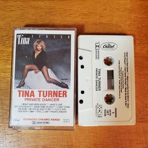 May include: A vintage cassette tape of Tina Turner's album "Private Dancer." The album cover features a photo of Tina Turner in a black outfit. The cassette tape is white with the album title and tracklist printed on it.