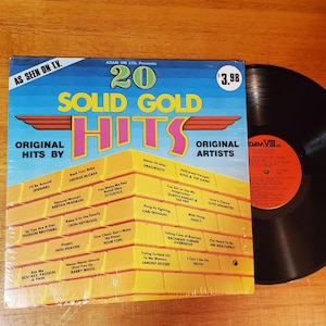 May include: A vinyl record album titled "20 Solid Gold Hits" with a blue, yellow, and pink design. The album cover features a list of song titles and artist names. The album is next to a black vinyl record with an orange label.