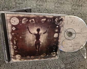Può includere: Un'immagine di un CD con una copertina color seppia che mostra una figura stilizzata con le braccia alzate, circondata da quadranti di orologi e altri simboli. Il CD è visibile accanto alla custodia. Il testo sul dorso recita "Ministry, K.E. & Ramsey, Sire/Warner Bros."