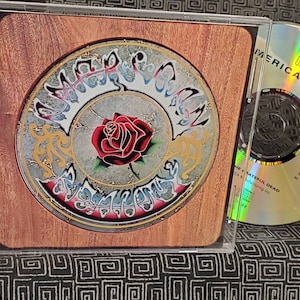 May include: A CD case with a wood-grain design featuring a circular image of a rose with a gold and blue design. The text "GRATEFUL DEAD/AMERICAN BEAUTY WARNER BROS." is printed on the side of the case. The CD is visible in the case.