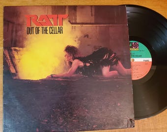 May include: A vintage vinyl record album by Ratt, titled "Out of the Cellar". The cover displays the band's red logo and a woman in a black outfit. A black vinyl record is partially visible beside the album cover.
