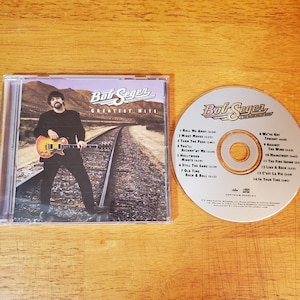May include: A Bob Seger Greatest Hits CD in a clear plastic case next to the disc. The cover shows a person holding a guitar on railroad tracks. The disc lists 14 tracks including Roll Me Away, Night Moves, and Turn the Page with Capitol Records text.