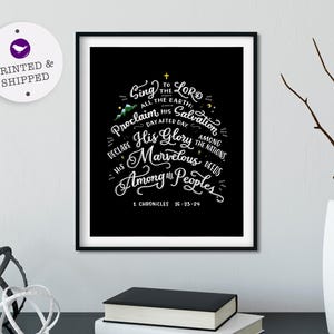 May include: Black framed art print with white script text. The text reads "Sing to the Lord all the earth...His Glory Marvelous Among All Peoples" from 1 Chronicles 16:23-24. A white vase with branches and a stack of books are in the foreground.