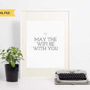 May include: A white framed print with the text "May the Wifi be with you" and a wifi symbol in grey. The print is sitting on a white surface with a potted plant and a vintage typewriter.