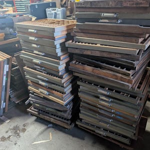 May include: A collection of vintage wooden printer's type cases, stacked in piles. The cases are primarily brown and gray, with metal handles and dividers. The image suggests a workshop or storage area, with other items visible in the background.
