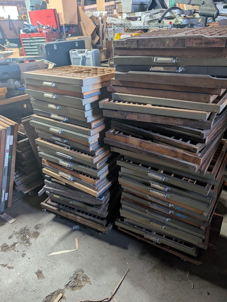 May include: A collection of vintage wooden printer's type cases, stacked in piles. The cases are primarily brown and gray, with metal handles and dividers. The image suggests a workshop or storage area, with other items visible in the background.