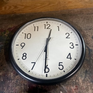May include: A round wall clock with a black frame and a white face featuring black Arabic numerals from 1 to 12. The clock has three black hands and the word ATOMIC printed in black below the center point, along with a small radio wave icon.